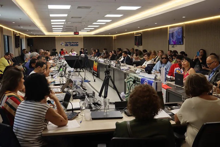 Imagem capta a grande mesa do plenario do CNS, em formato retangular, com todos os conselheiros sentados. Ao fundo, logomarca do CNS.