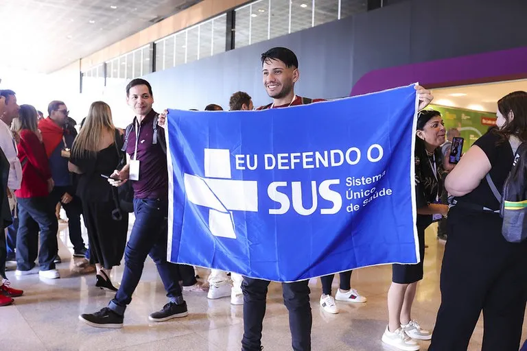 Foto de participante da 5ª Conferência Nacional de Saúde do Trabalhador e da Trabalhadora, realizada em agosto de 2025. Em pé, ele segura uma grande bandeira azul, onde está escrito: Eu Defendo o SUS.
